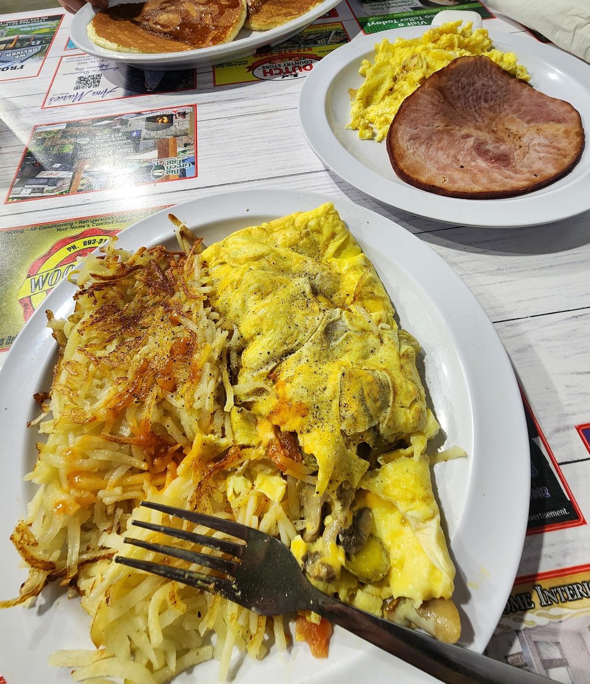 An omelette with hash browns, a plate of ham steak and scrambled eggs, and a plate of pancakes across a diner table.